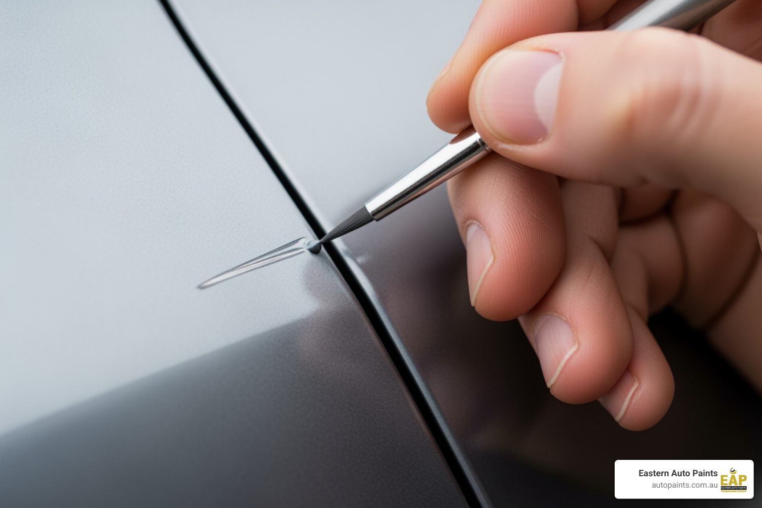 Close-up of a hand using a fine-tipped detail brush for automotive touch up on a silver car panel scratch.
